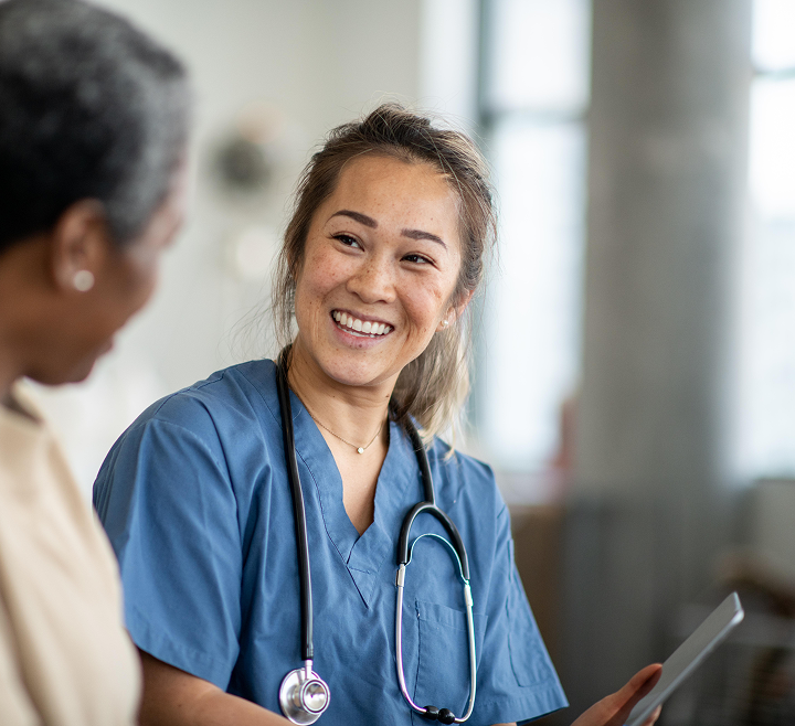 Smiling nurse in blue scrubs with a stethoscope, speaking with a patient in a bright healthcare setting.